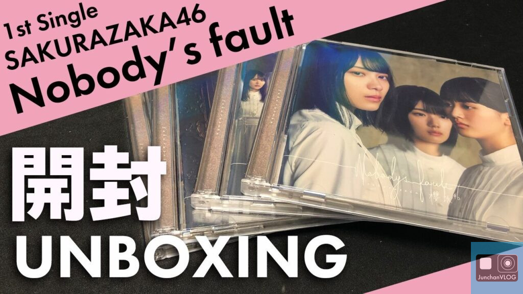 白い服を着た3人の人物が描かれたカバーアートのCDが積み重ねられています。一番上のCDは黒い表面に斜めに置かれています。大きなフォントで「1st Single SAKURAZAKA46 Nobodys fall」と「開封 UNBOXING」と書かれています。.