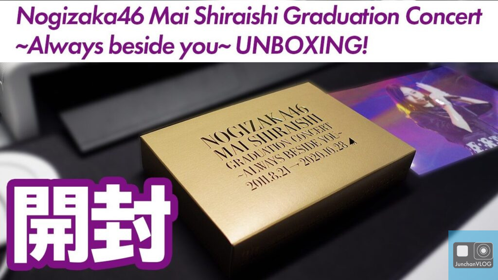 黒い表面に「乃木坂46 白石麻衣卒業コンサート ~Always beside you~」と書かれた金色の箱。背景にはステージで演奏している人物のぼやけた映像が描かれている。上部には紫と白の大きな文字で「UNBOXING!」と書かれている。.