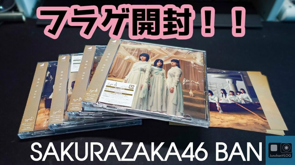 同じ明るい色のドレスを着た3人の女性が描かれたCDジュエルケースの山。右に同じグループの写真が置かれている。暗い背景に太字の日本語と英語のテキストで「フラゲ開封!! SAKURAZAKA46 BAN」と書かれている。.