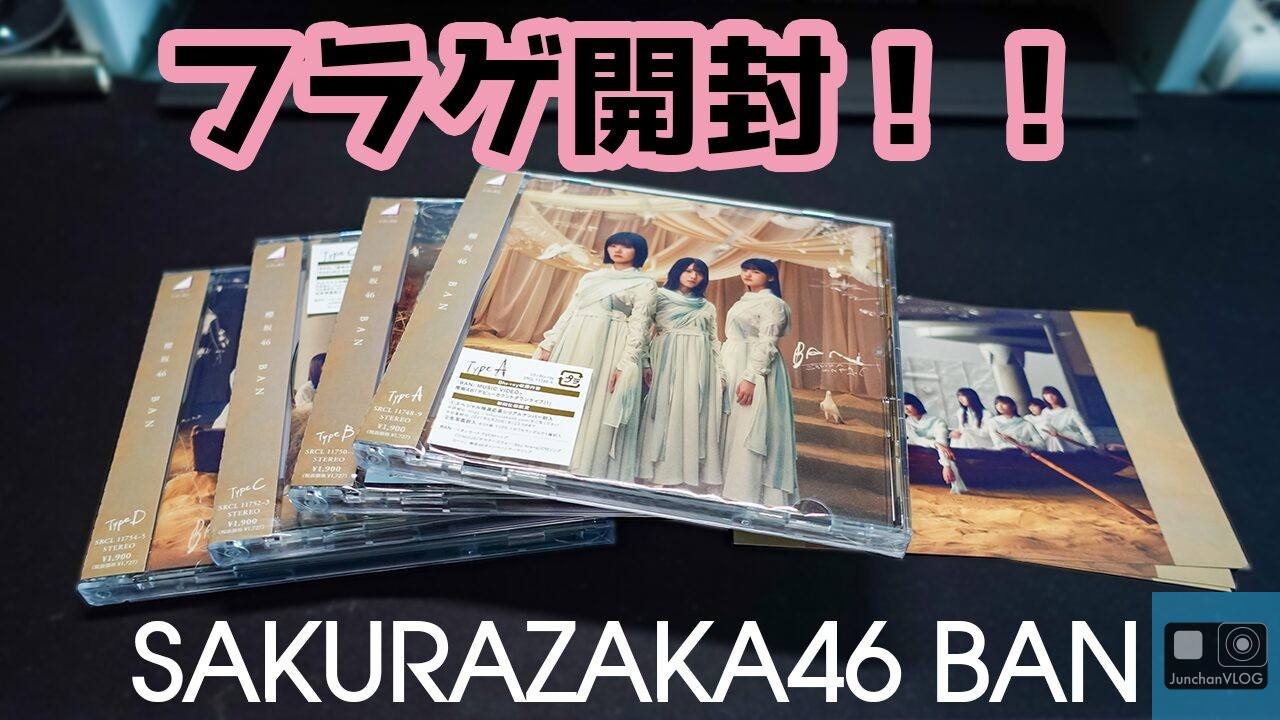 同じ明るい色のドレスを着た3人の女性が描かれたCDジュエルケースの山。右に同じグループの写真が置かれている。暗い背景に太字の日本語と英語のテキストで「フラゲ開封!! SAKURAZAKA46 BAN」と書かれている。.